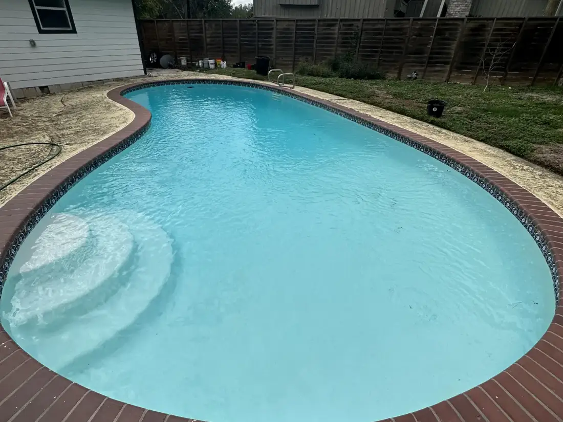 The same kidney pool after replastering: clear blue water over new white plaster, same house and fence behind